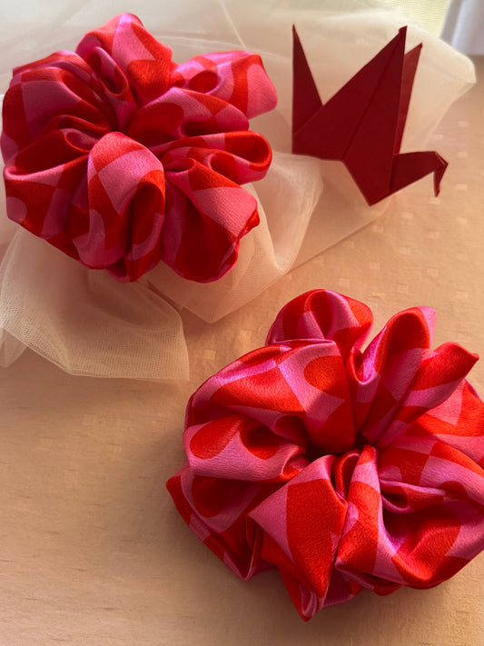 Two XL satin scrunchies in pink with cute heart patterns, designed for all hair types (thick, thin, or voluminous) as a gentle yet secure hair tie or trendy wrist accessory.