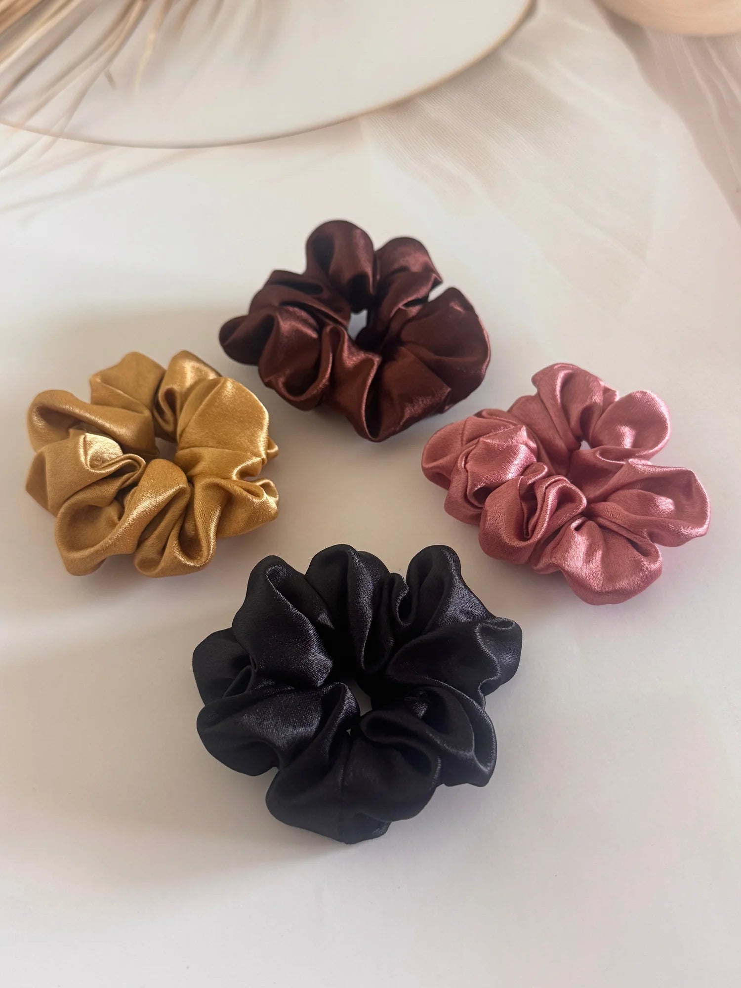 Four satin scrunchies in black, gold, rust, and burgundy displayed on white surface by Bows and Bags India