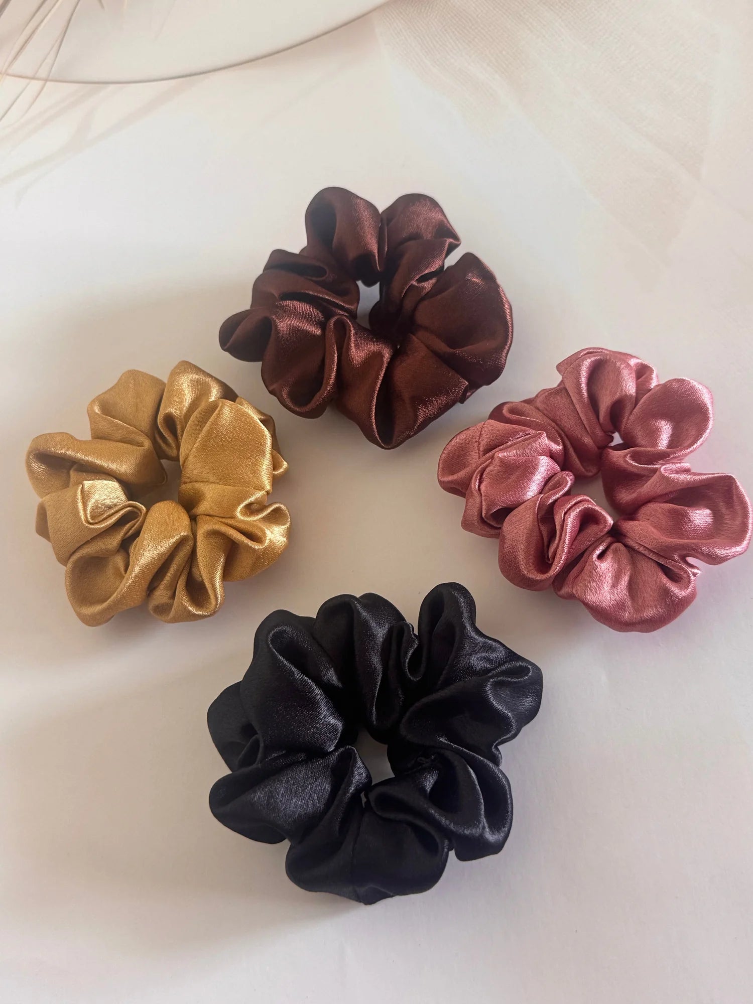 Four premium satin scrunchies in black, gold, rust, and burgundy by Bows and Bags India displayed flat lay on white fabric