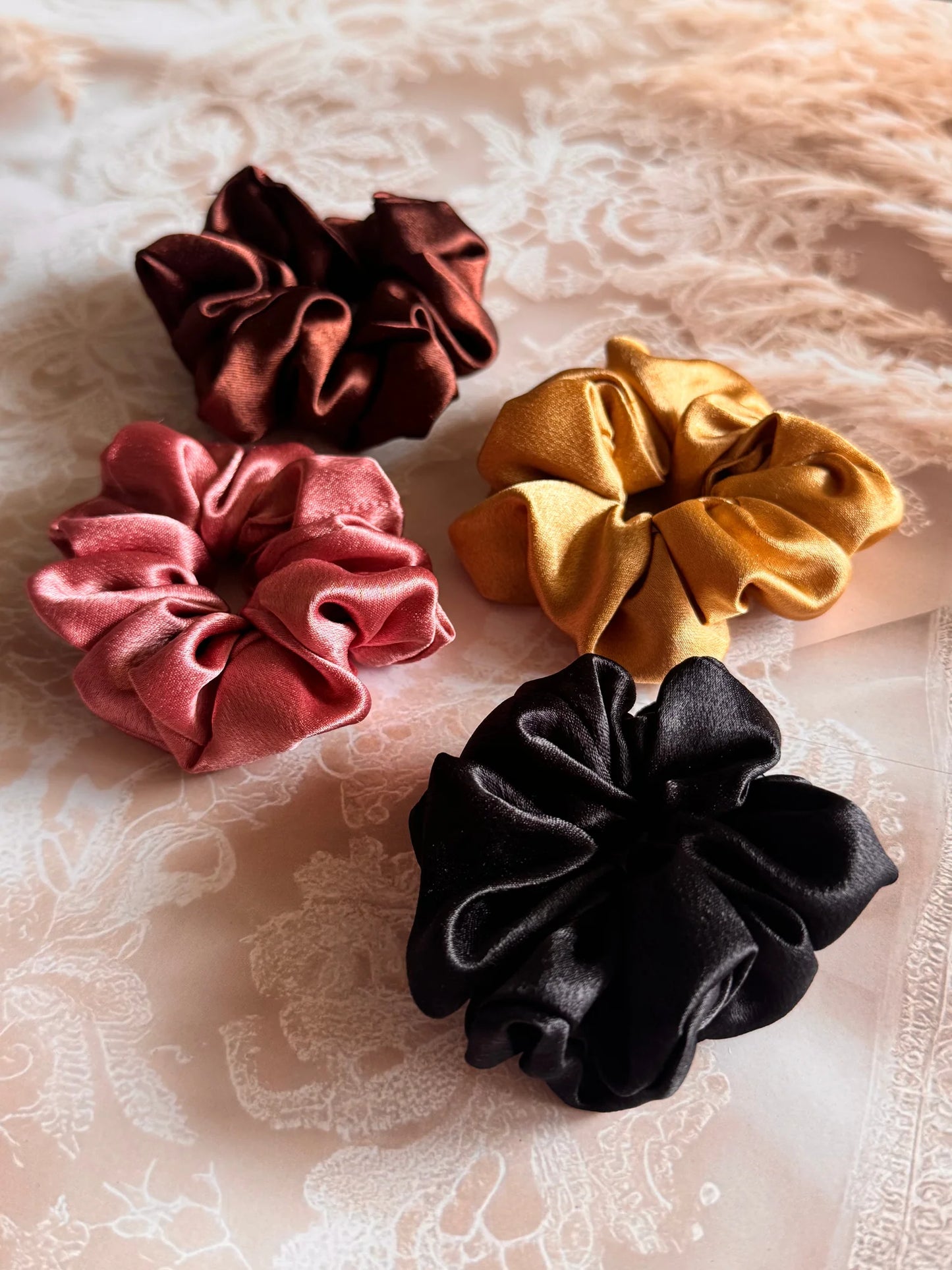 Four premium satin scrunchies in black, gold, rust, and burgundy by Bows and Bags India displayed on white lace fabric