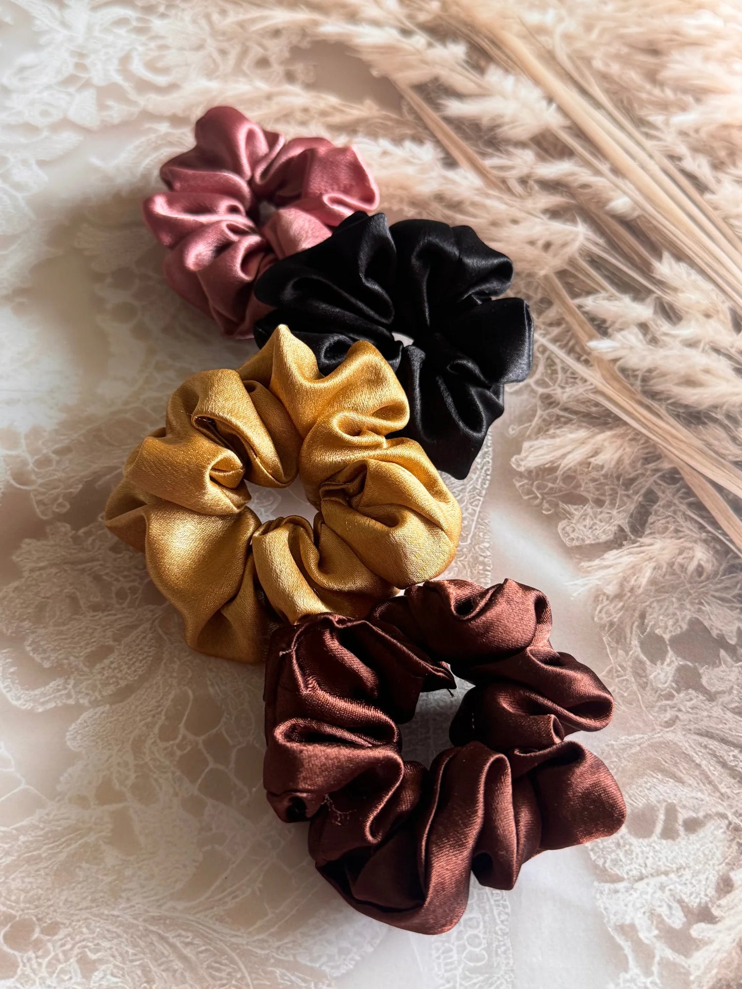 Four premium satin scrunchies in black, gold, rust, and burgundy by Bows and Bags India, displayed on textured fabric