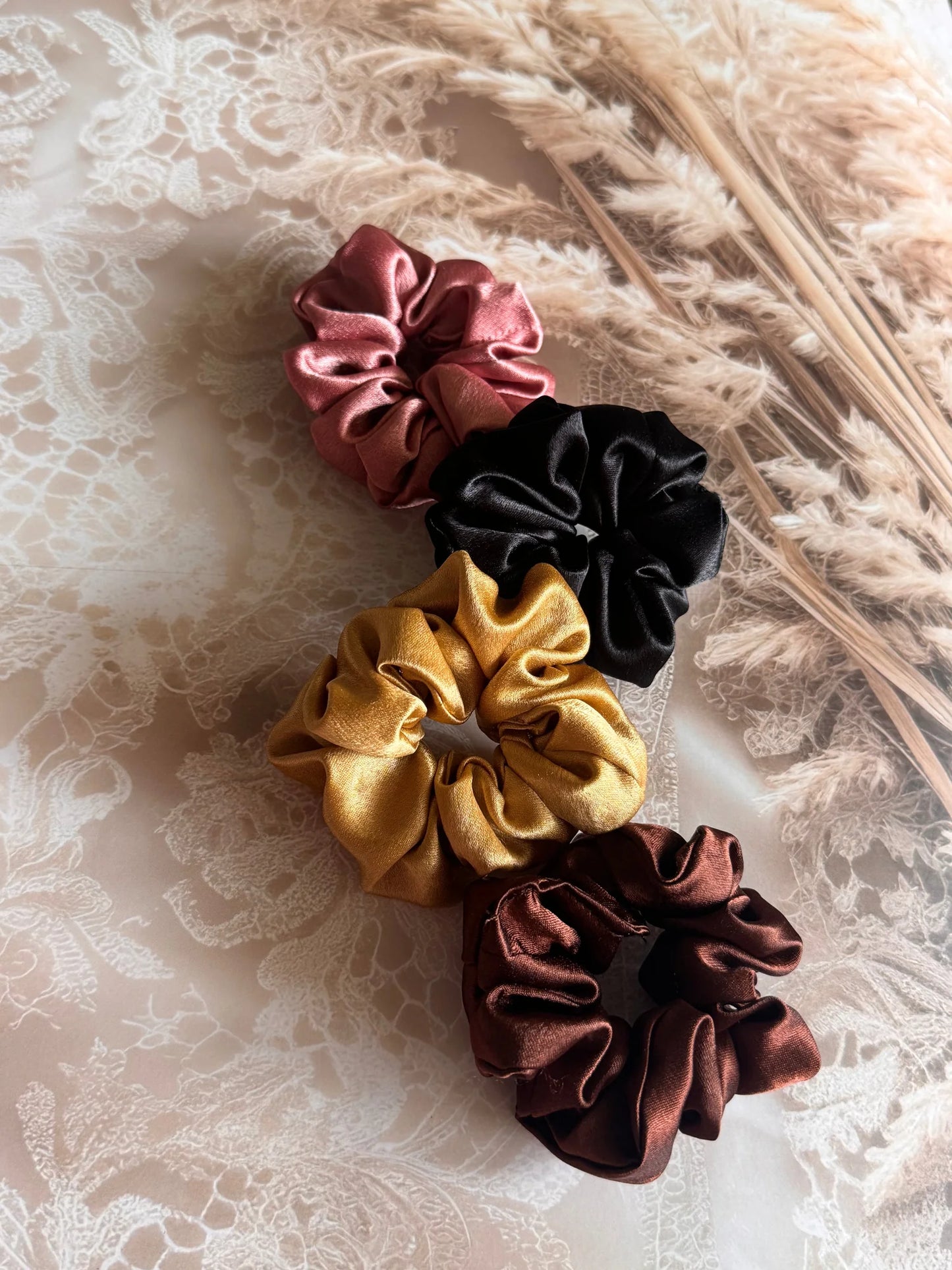 Four satin scrunchies in black, gold, rust, and burgundy by Bows and Bags India, flat lay on lace background showing