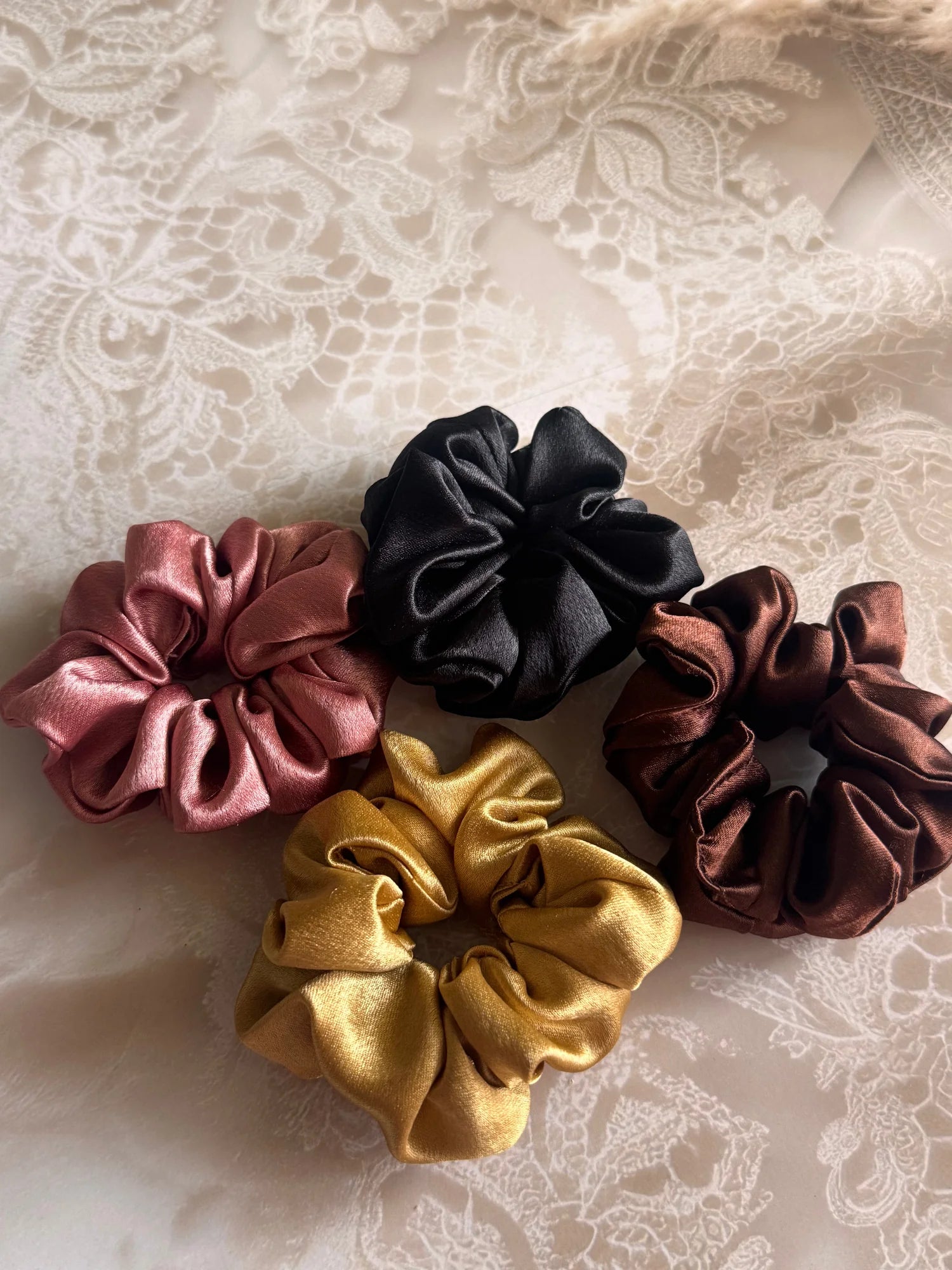 Four premium satin scrunchies in black, gold, rust, and burgundy by Bows and Bags India, displayed on lace background
