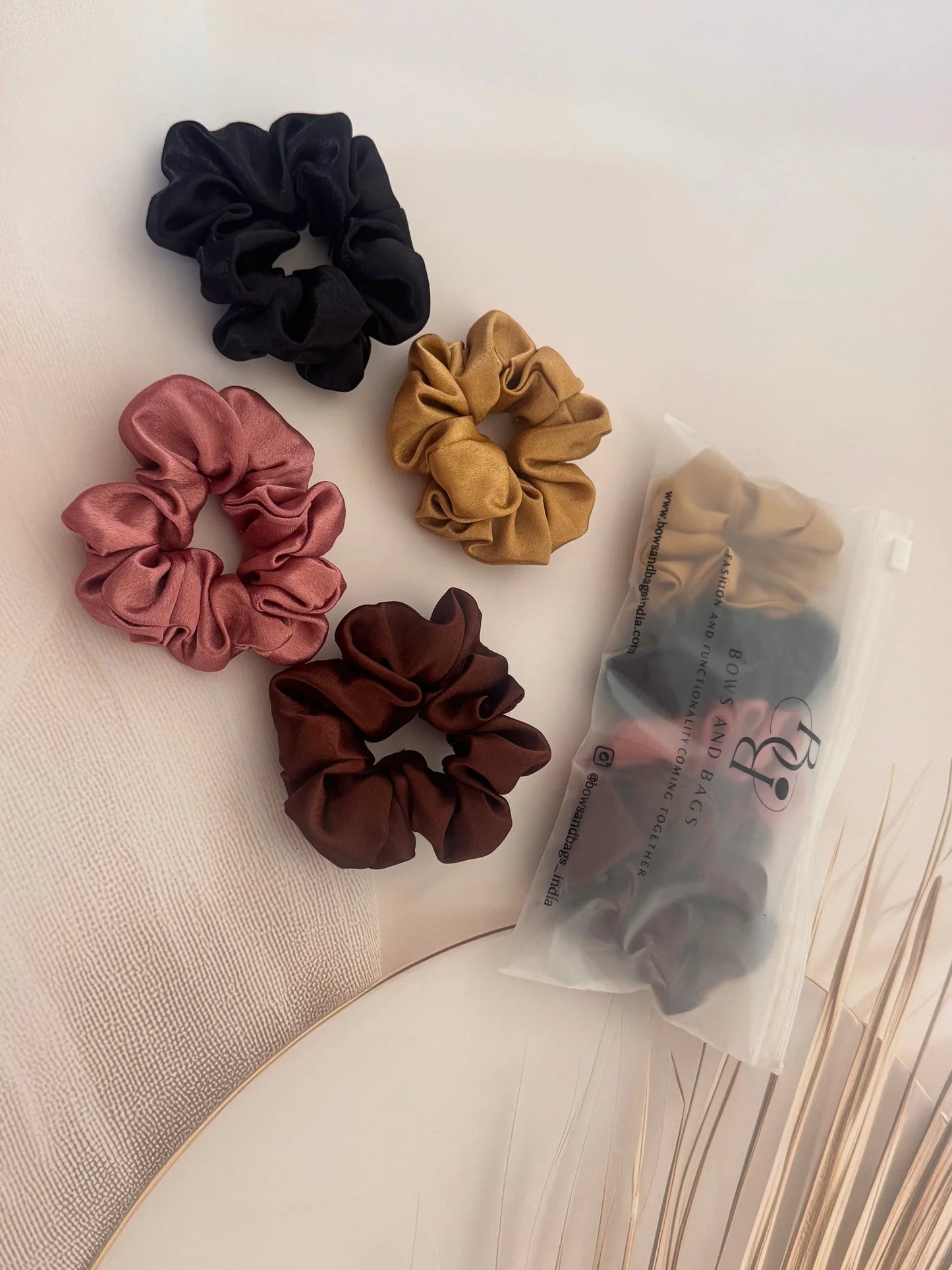 Four satin scrunchies in black, gold, rust, and burgundy colors displayed with packaging by Bows and Bags India handmade