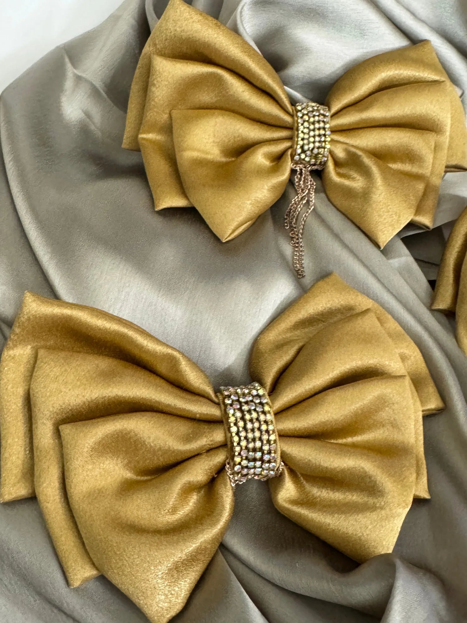 Two gold satin hair bows with stone and chain embellishments by Bows and Bags India, displayed on neutral fabric backdrop