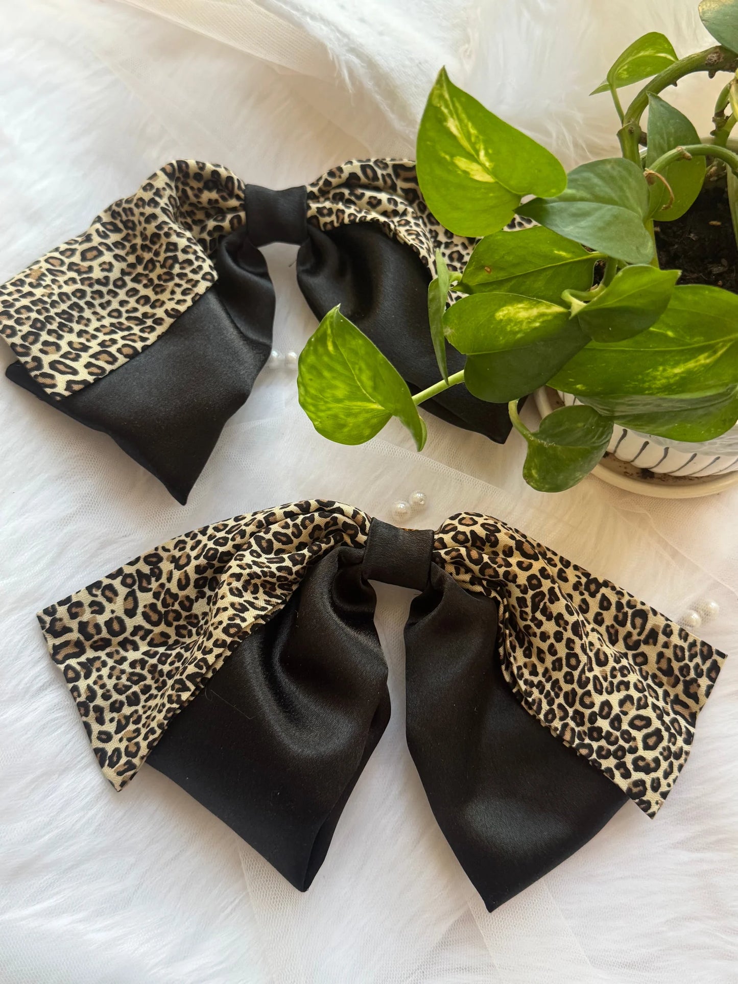Two handmade Kate Moss hair bows featuring black satin with leopard print, displayed with green plant on white surface by