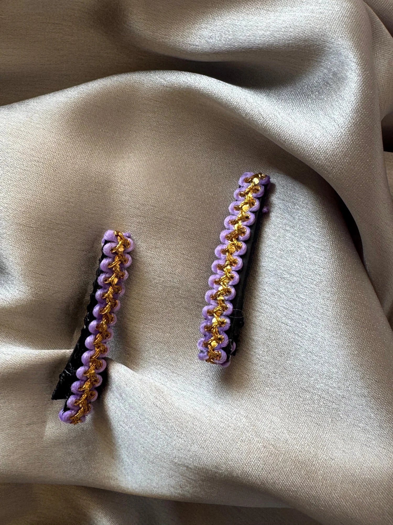 Pair of lavender and gold handmade hair clips by Bows and Bags India displayed on neutral beige fabric, showing intricate