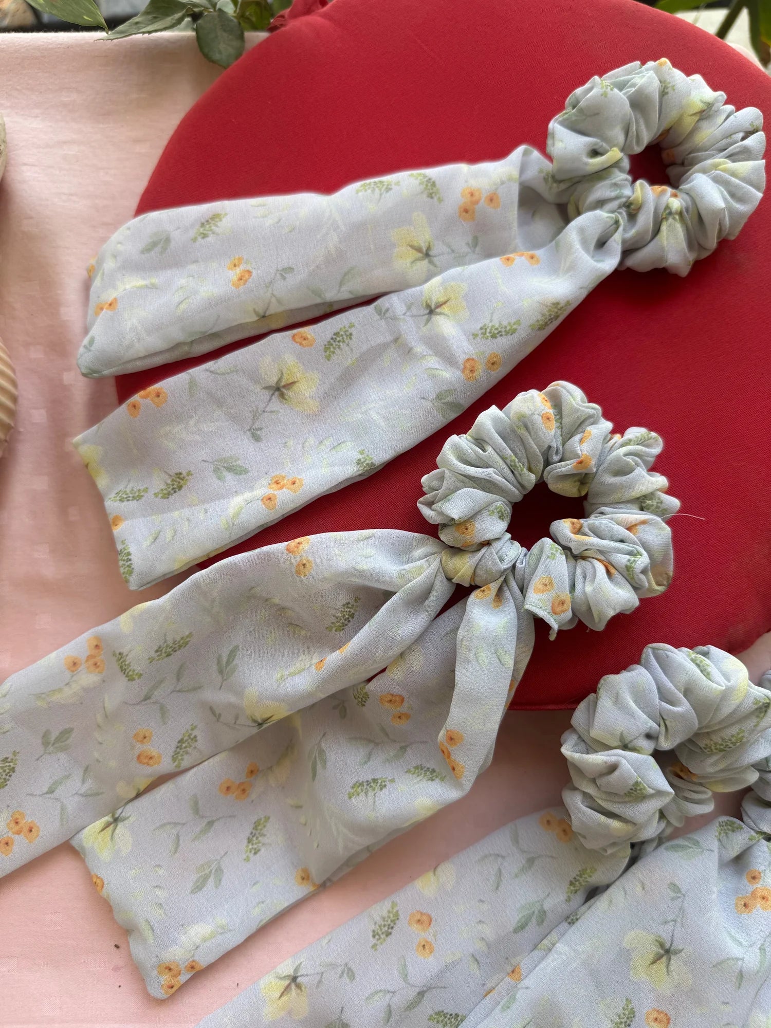 Light green floral georgette tail scrunchies with gathered fabric ends by Bows and Bags India, displayed on red and pink