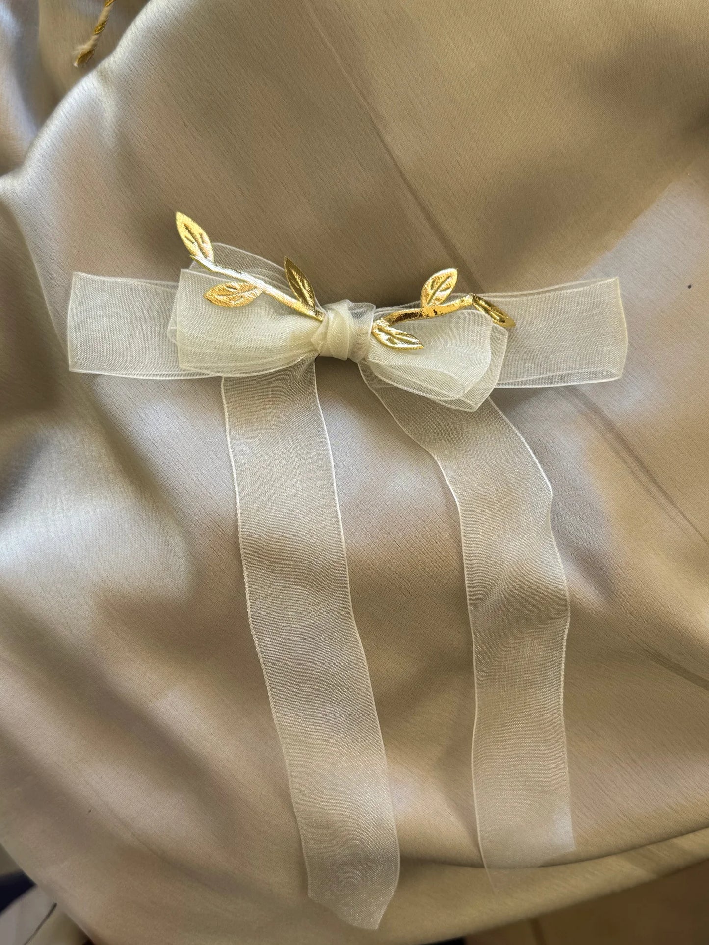 White sheer organza bow hair clip with gold leaf accents by Bows and Bags India, close-up detail view on cream fabric