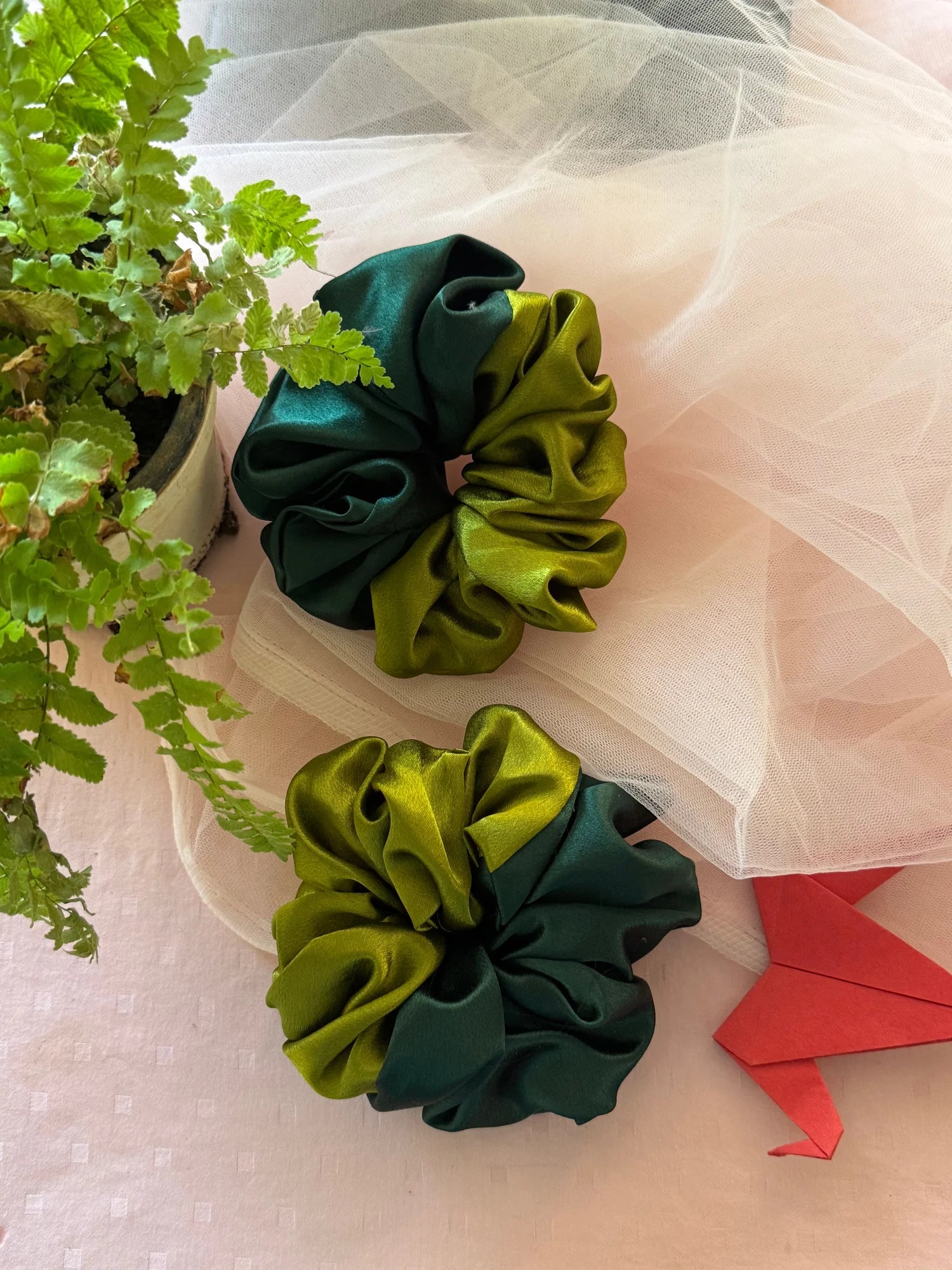 Olive Garden dual-tone satin scrunchies in olive and dark green displayed on sheer tulle fabric with fresh green foliage by