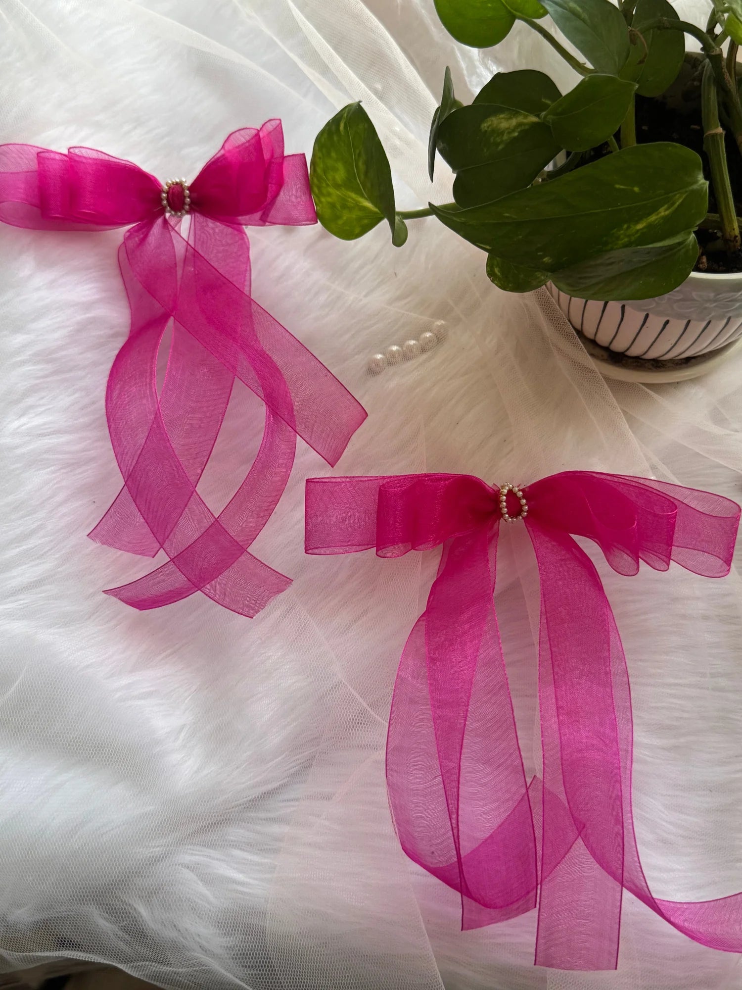 Hot pink organza hair bow clip by Bows and Bags India displayed on white fabric with green plant, showing sheer texture and