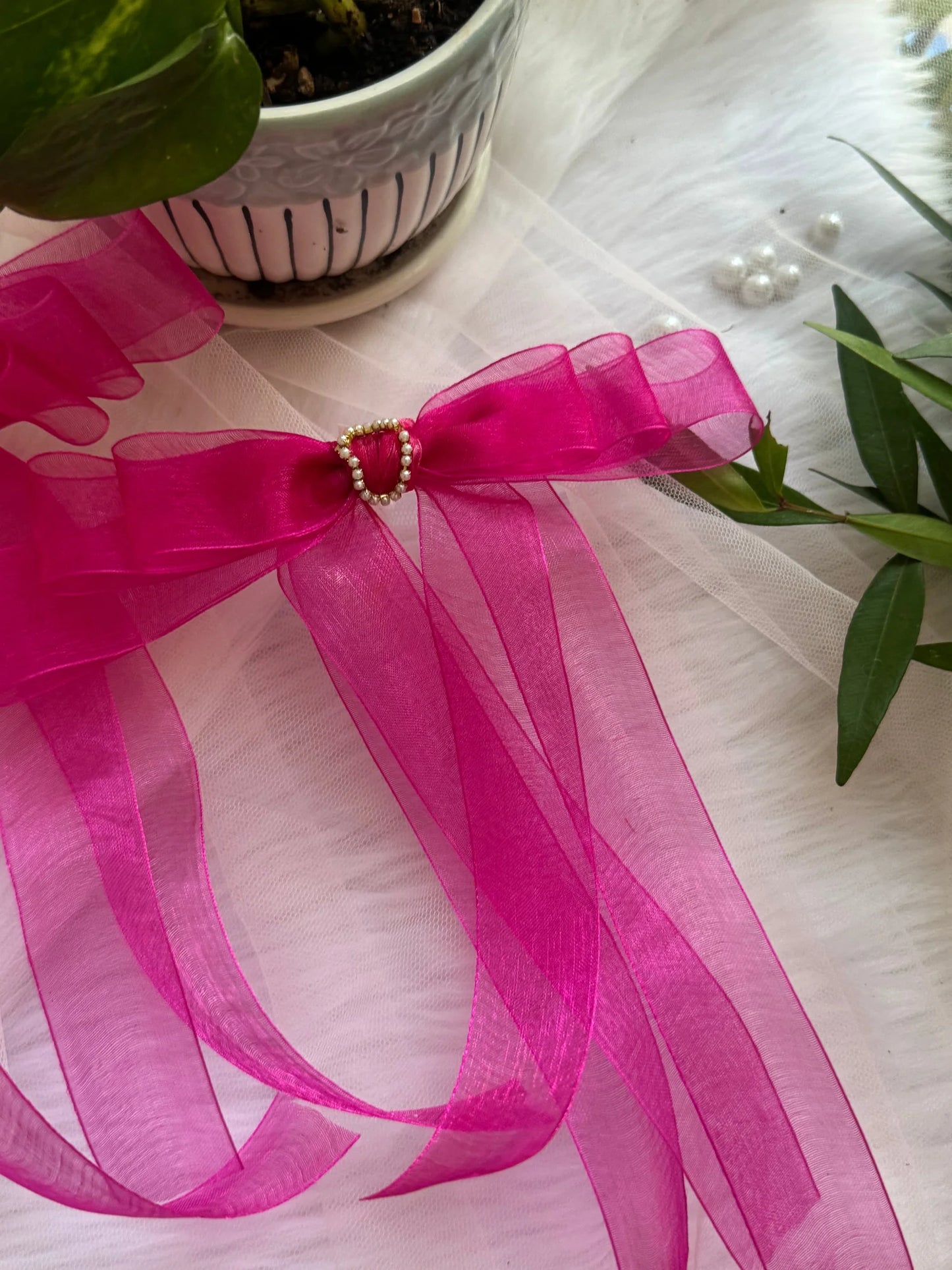 Pink organza hair bow with pearl bead center detail by Bows and Bags India, displayed on white fabric with green plant