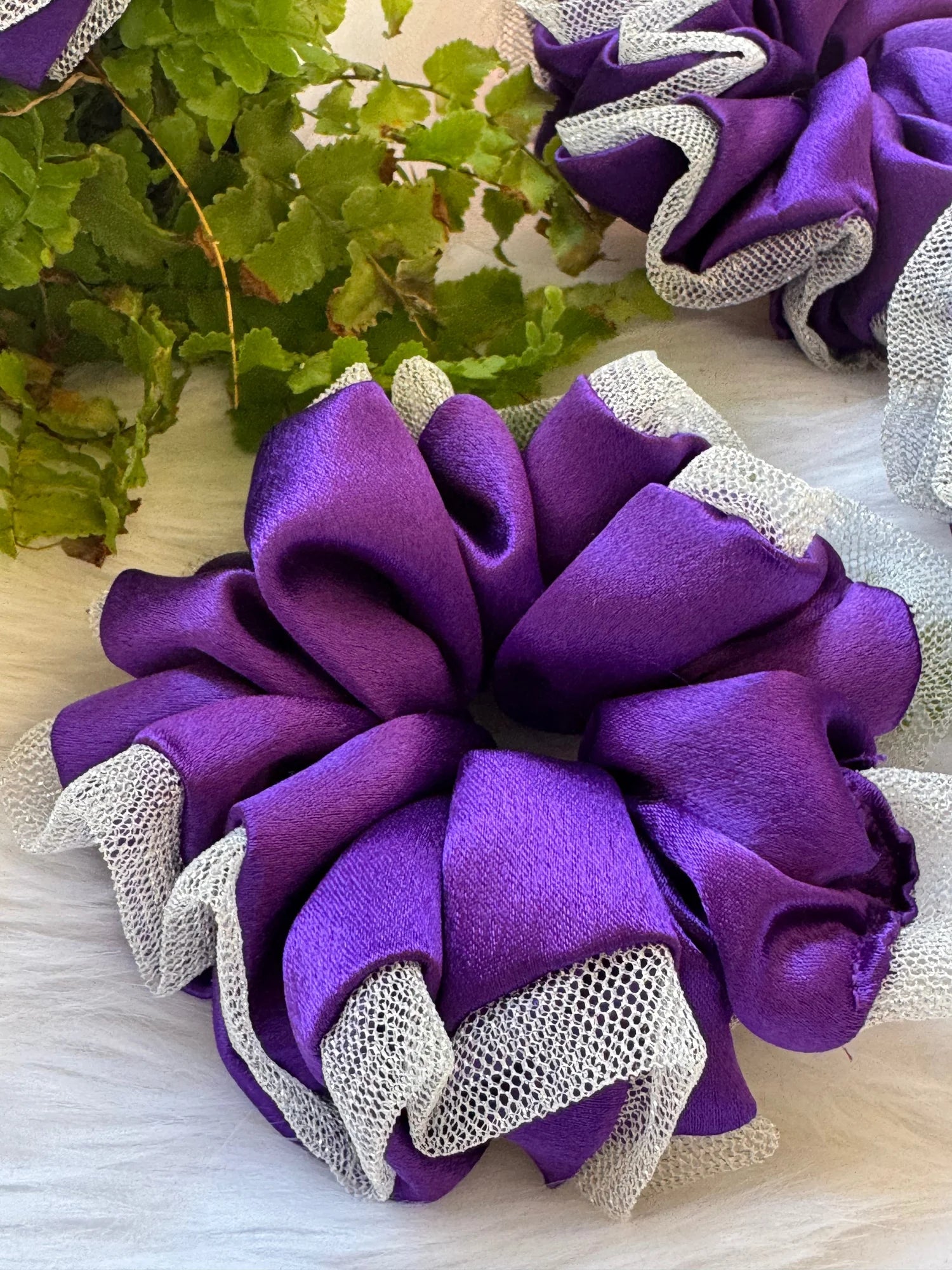 Purple satin scrunchie with silver net border by Bows and Bags India, handmade hair accessory displayed with green foliage