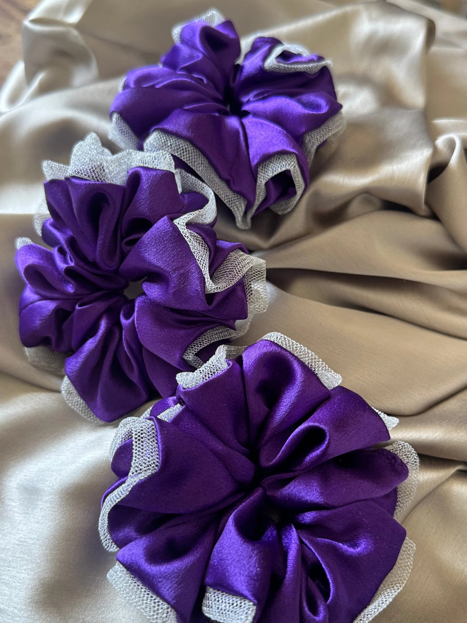 Three purple satin scrunchies with silver net borders by Bows and Bags India, displayed on cream fabric showing handmade