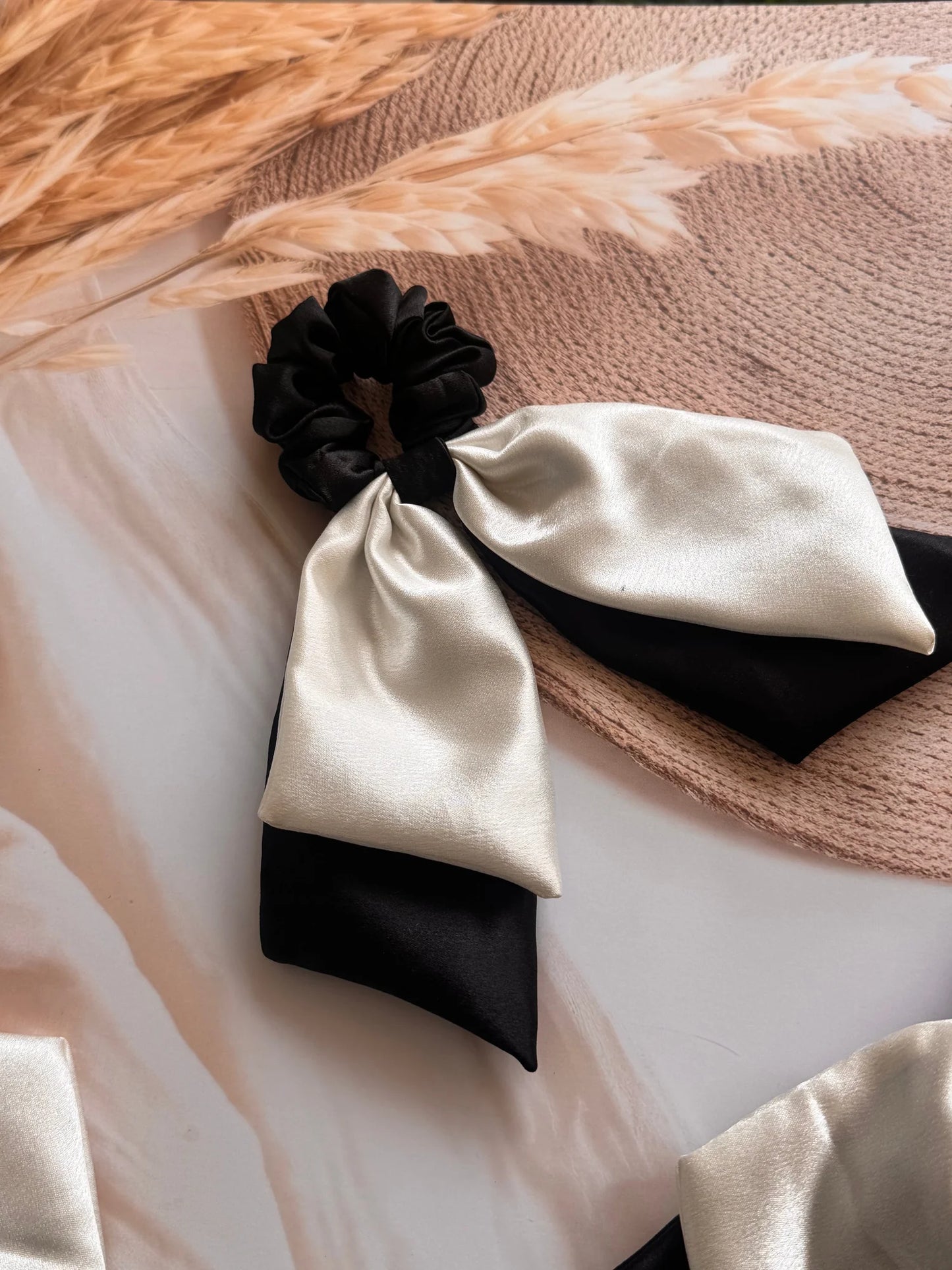 Black and white satin dual tail bow scrunchie by Bows and Bags India displayed on neutral textured fabric, showing handmade