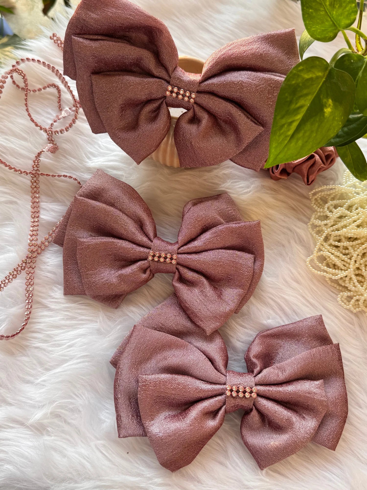 Three handmade rose gold silk hair bows with pearl detailing by Bows and Bags India, displayed on white fur with green