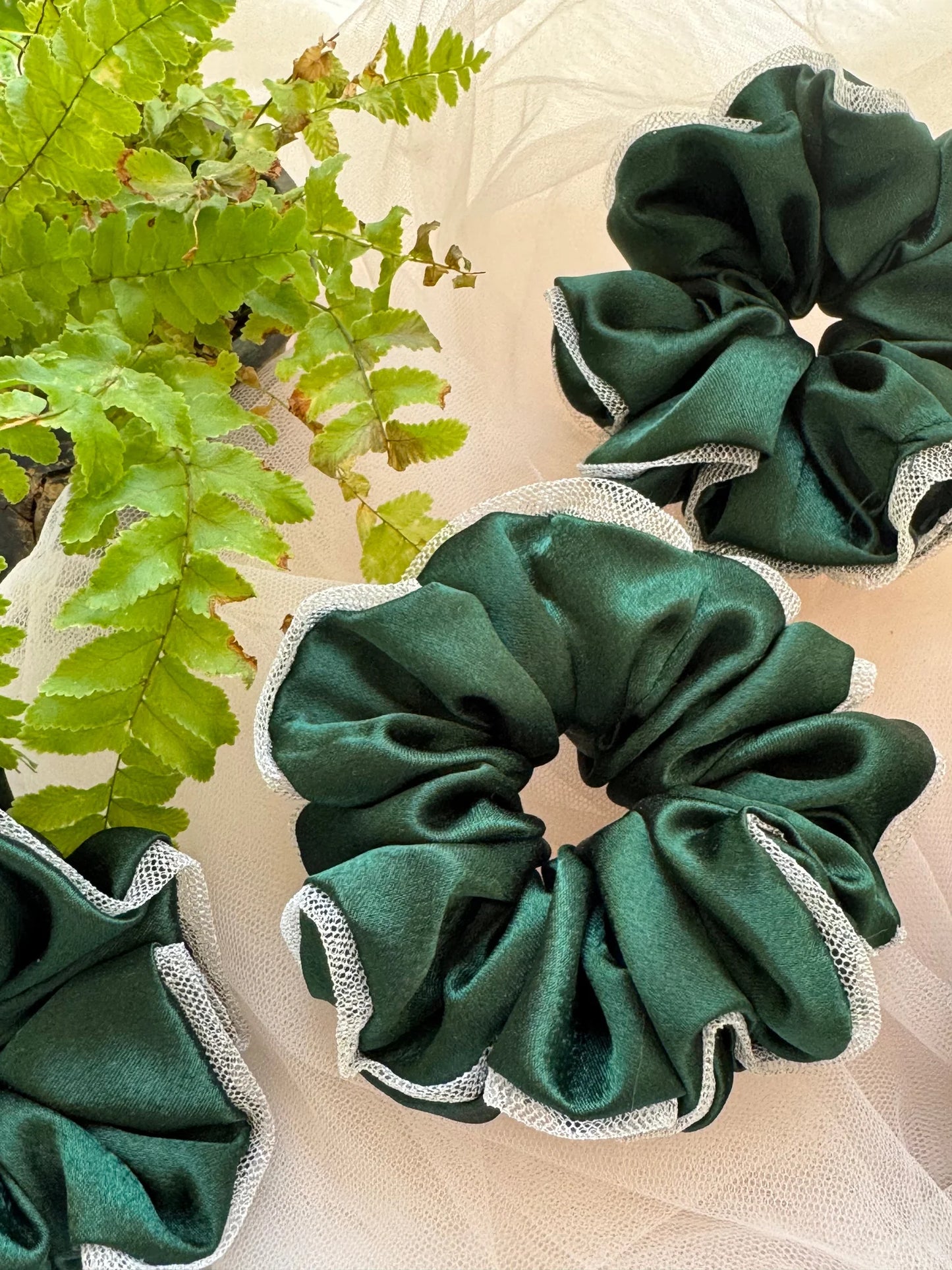 Dark green satin scrunchie with silver net border by Bows and Bags India, displayed with fresh green fern leaves on neutral