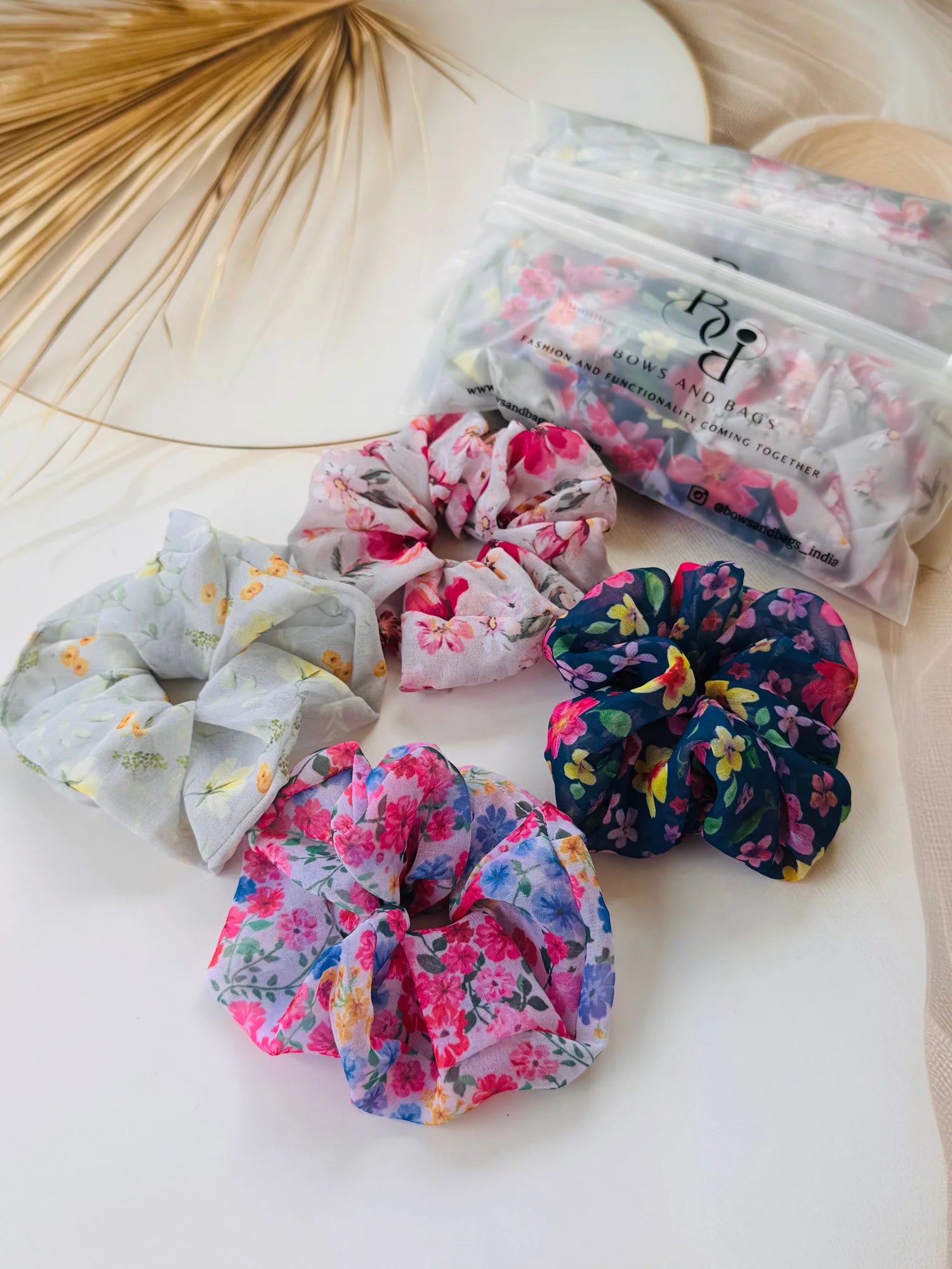 Four assorted floral georgette scrunchies from Bows and Bags India displayed with packaging, showing vibrant pink, blue, and