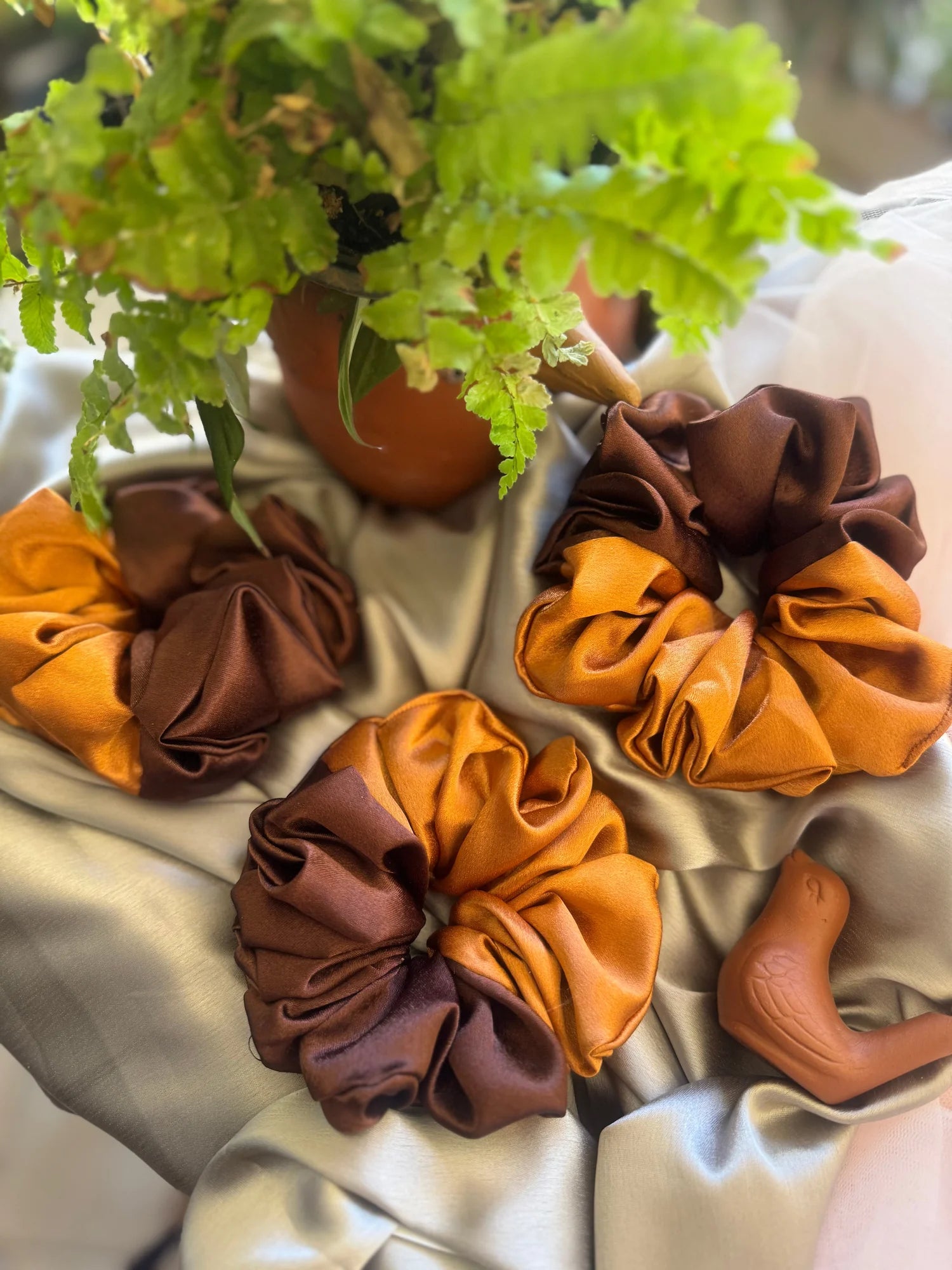 Three handmade satin scrunchies in terracotta and brown by Bows and Bags India, displayed on white fabric with green plant